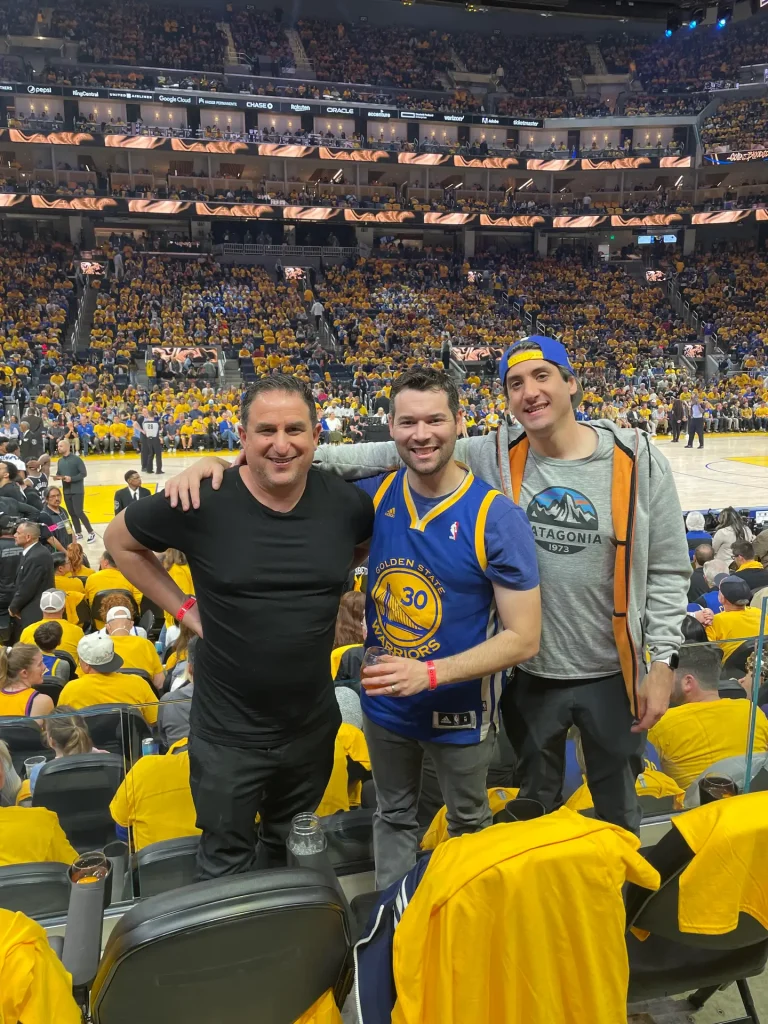 Three men enjoying a live basketball game wearing casual and custom sports apparel, showcasing corporate swag and branded event merchandise at a packed stadium.