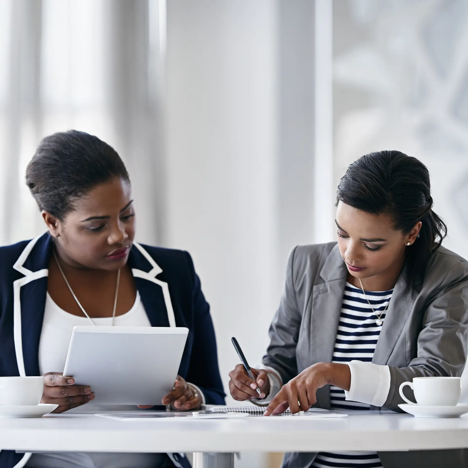 Two professional women collaborating in office meeting, reviewing documents and taking notes, corporate teamwork and business planning concept.
