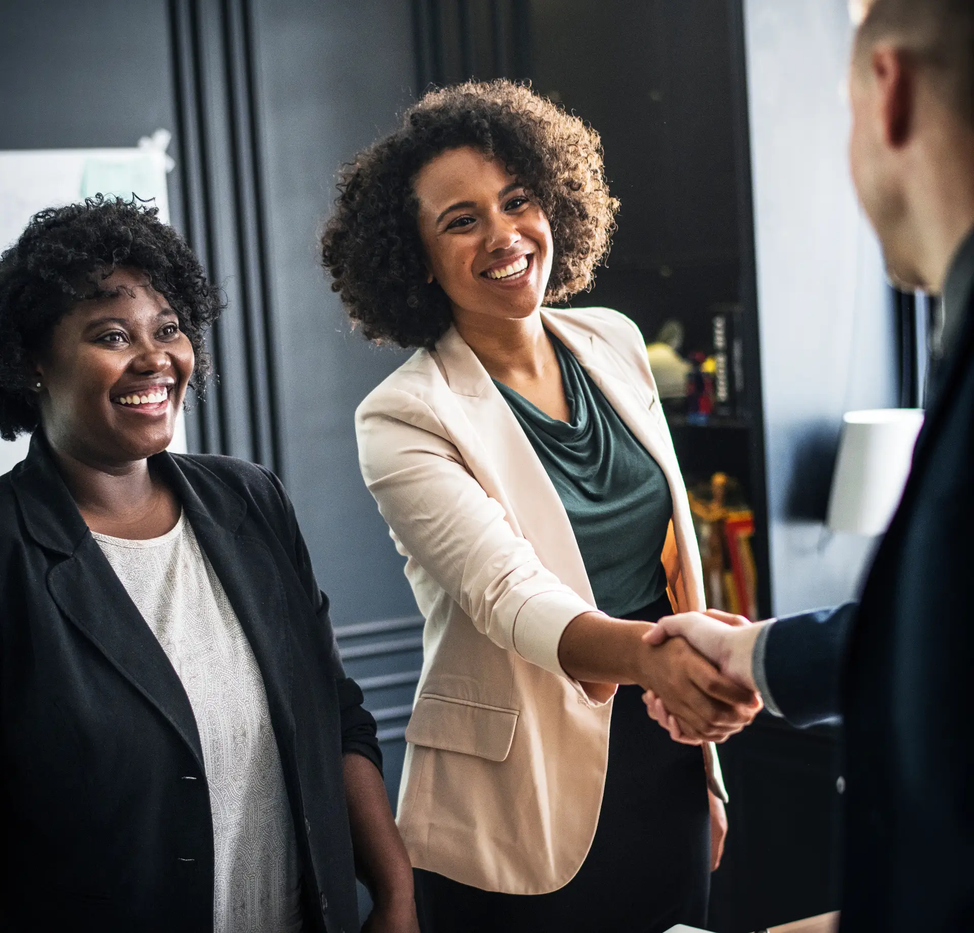 Two professional women smiling and shaking hands with a businessperson in a modern office, representing corporate client engagement and partnership with a top corporate swag vendor.