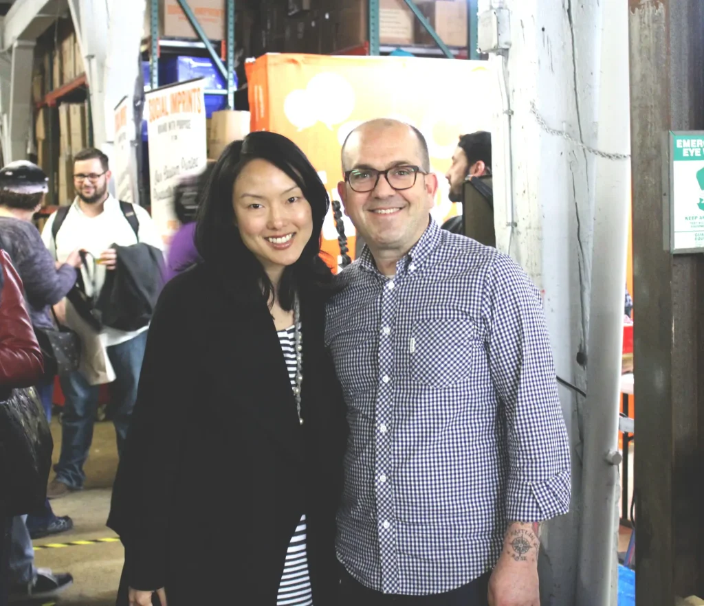 Two smiling professionals posing at a busy warehouse event for Social Imprints, a top corporate swag company specializing in custom promotional products, live screen printing, and sustainable swag solutions in San Francisco.