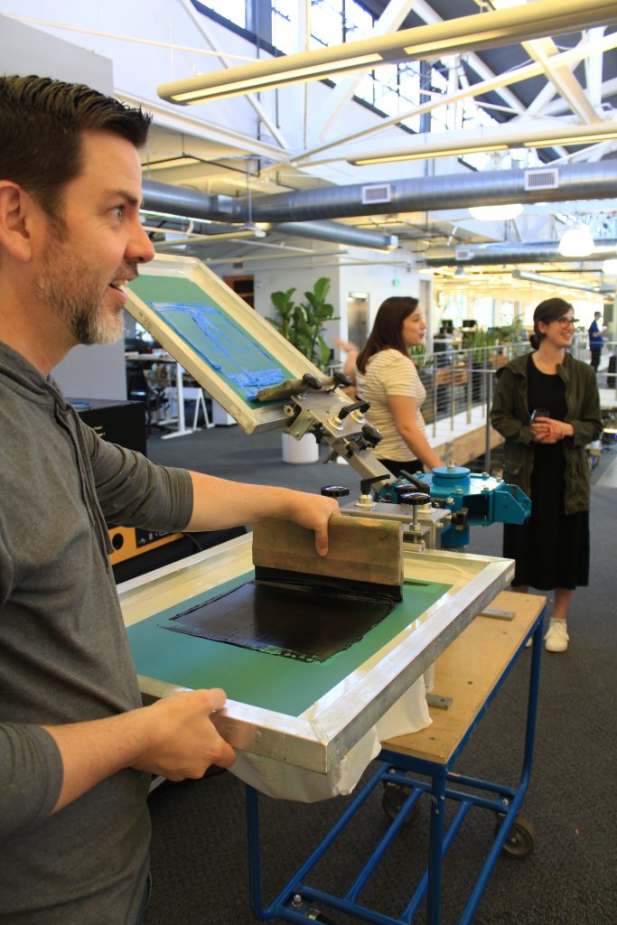 Man demonstrating live screen printing on custom promotional products in a modern office setting, showcasing corporate swag vendor services and event giveaway solutions.