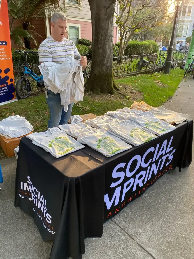 Man arranging custom printed eco-friendly promotional t-shirts on a branded Social Imprints table at an outdoor event, showcasing sustainable swag and corporate giveaway items for events in San Francisco.