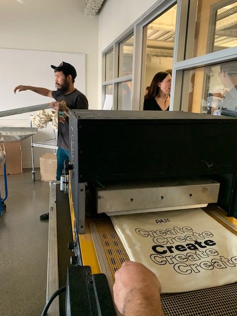 Live screen printing of custom branded tote bags at a professional promotional products company specializing in corporate swag, event giveaways, and sustainable branded merchandise in San Francisco.