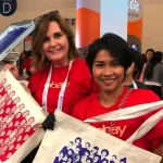 Two smiling women wearing red shirts showcasing custom printed tote bags at a live screen printing event, highlighting promotional products and branded swag for corporate events and giveaways.