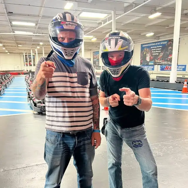 Two men wearing helmets and casual clothing pose at an indoor go-kart racing track, showcasing team-building and corporate event fun.