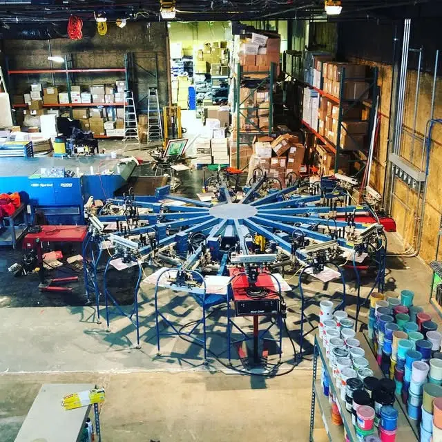 Industrial screen-printing machine in a spacious warehouse for custom branded merchandise production, showcasing live printing and fulfillment services for corporate swag and promotional products in San Francisco.
