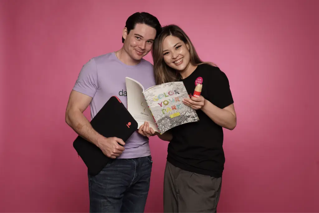 Two smiling professionals holding custom branded promotional products including a laptop sleeve and a colorful branded booklet, showcasing corporate swag and employee gifts against a pink background.