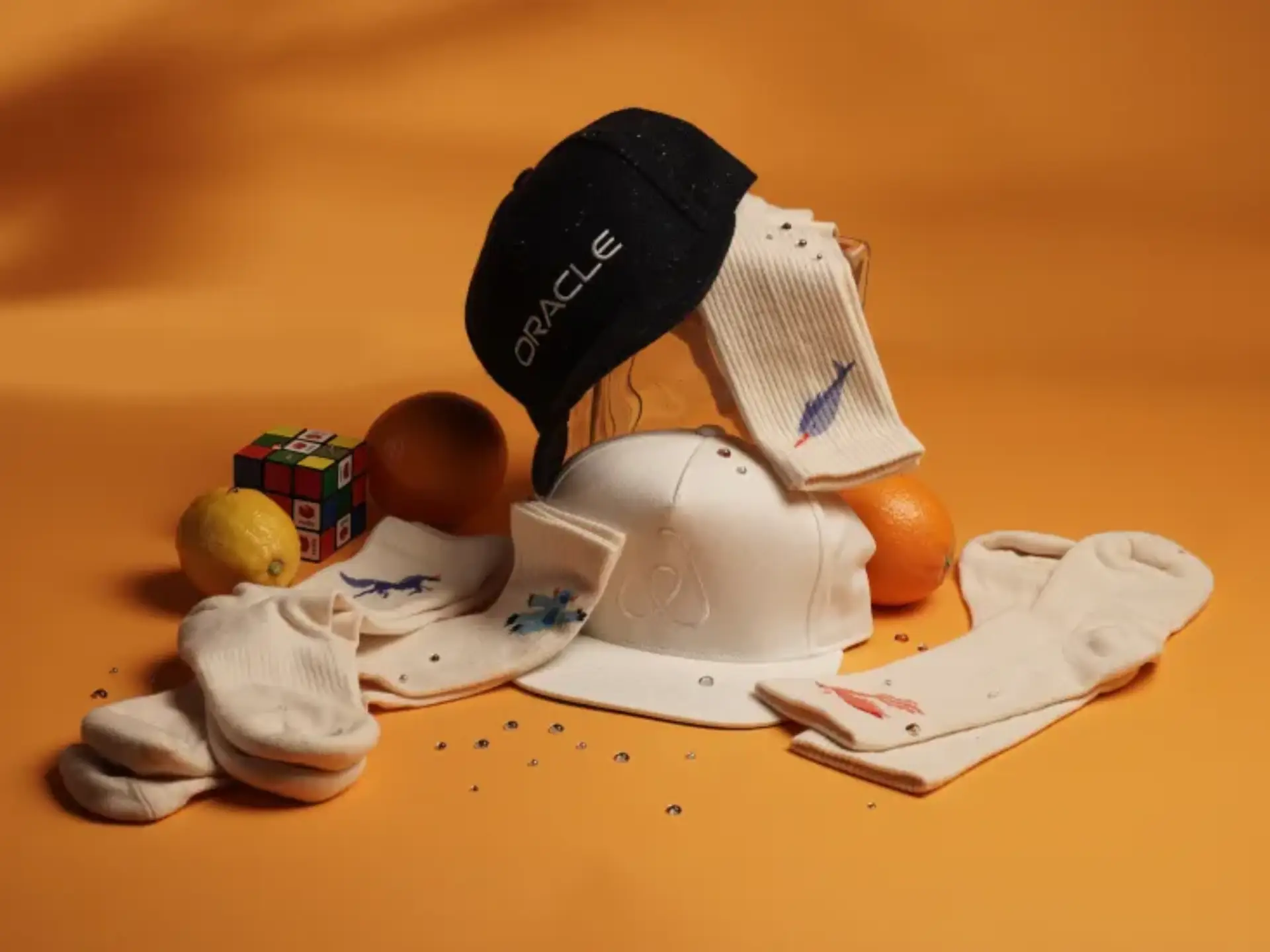 Custom branded promotional socks and hats with embroidered logos, styled with fruit and a Rubikâ€™s cube on an orange background, showcasing eco-friendly corporate swag and event giveaway items.