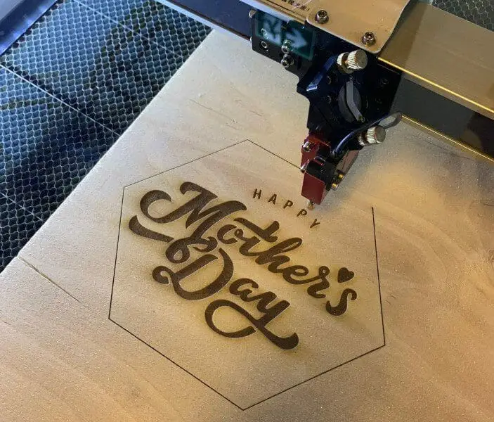Laser engraving machine creating a custom "Happy Mother's Day" wooden plaque, showcasing personalized promotional products and custom branded merchandise for corporate gifts and employee recognition.