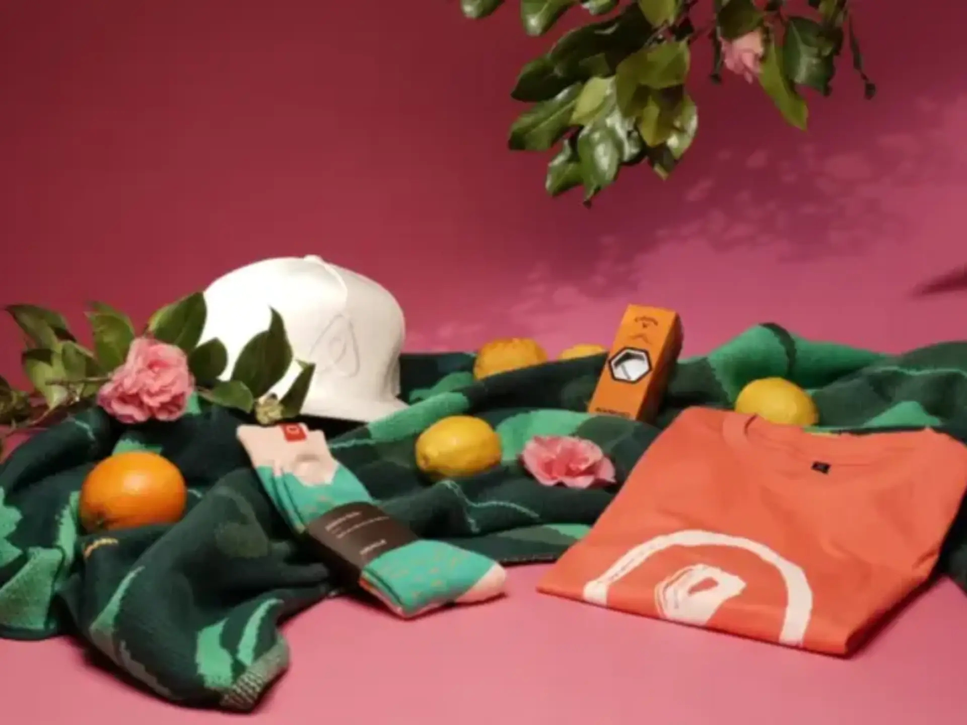 Branded corporate swag including custom printed t-shirt, white cap, colorful socks, and eco-friendly promotional products styled with fresh citrus and flowers for sustainable event giveaways.