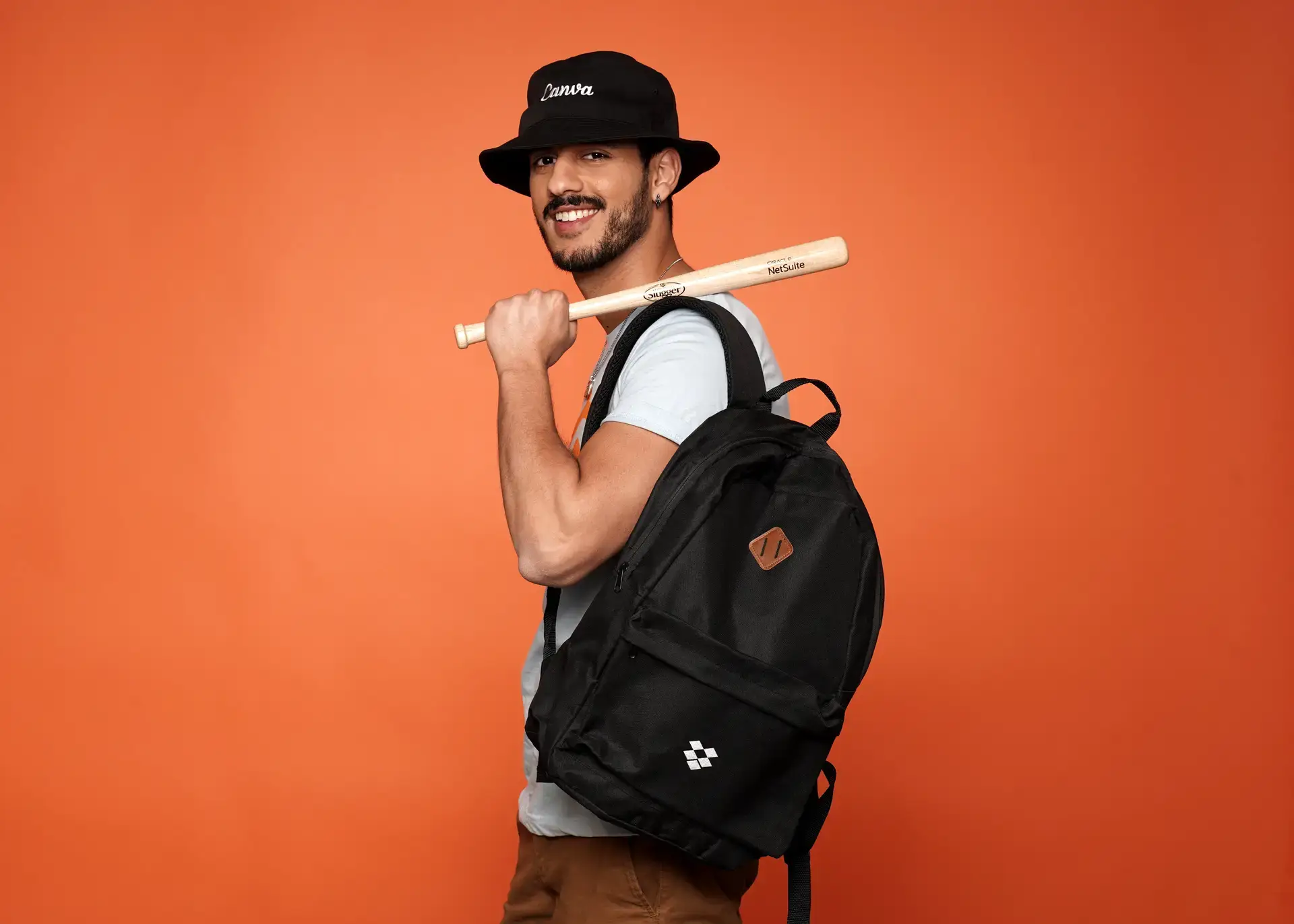 Smiling man wearing custom branded black backpack and bucket hat holding personalized promotional baseball bat against orange background, showcasing corporate swag and event giveaway items.