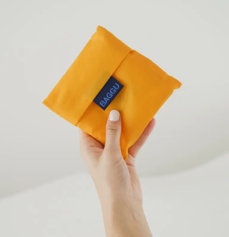 Hand holding compact folded orange reusable BAGGU bag, eco-friendly promotional product ideal for sustainable swag and corporate giveaways.