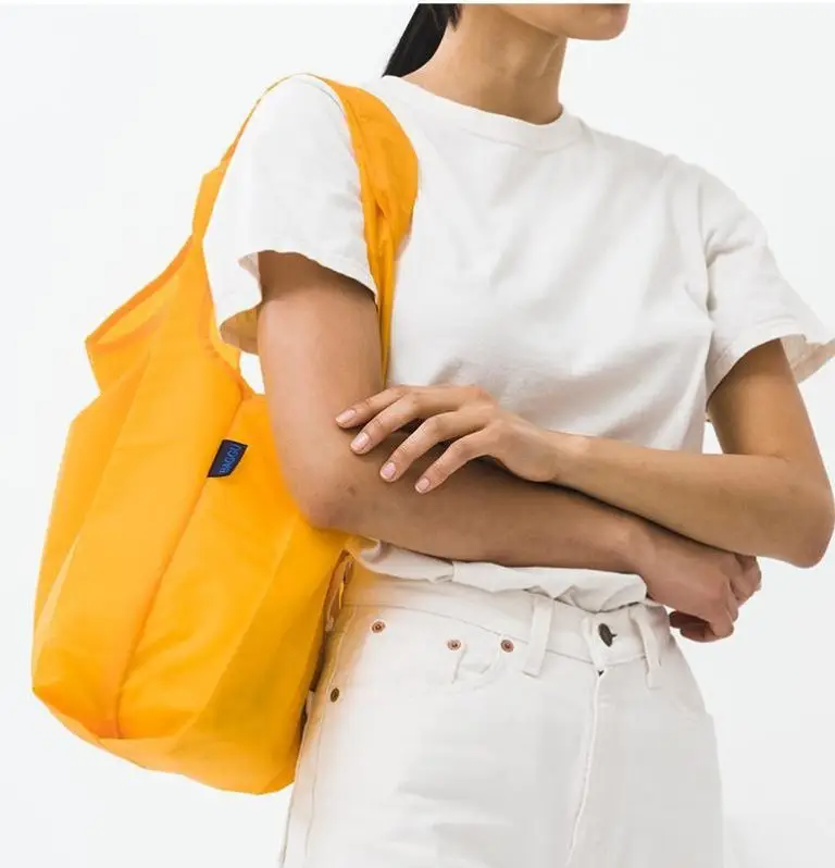 Woman wearing white tee and white pants carrying bright yellow reusable tote bag, eco-friendly promotional swag item for corporate events and giveaways.